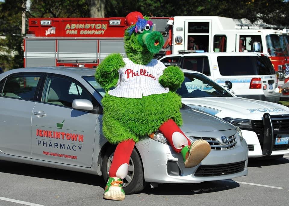 The Phillie Phanatic leaning on a Jenkintown Pharmacy free same-day delivery car at a community event